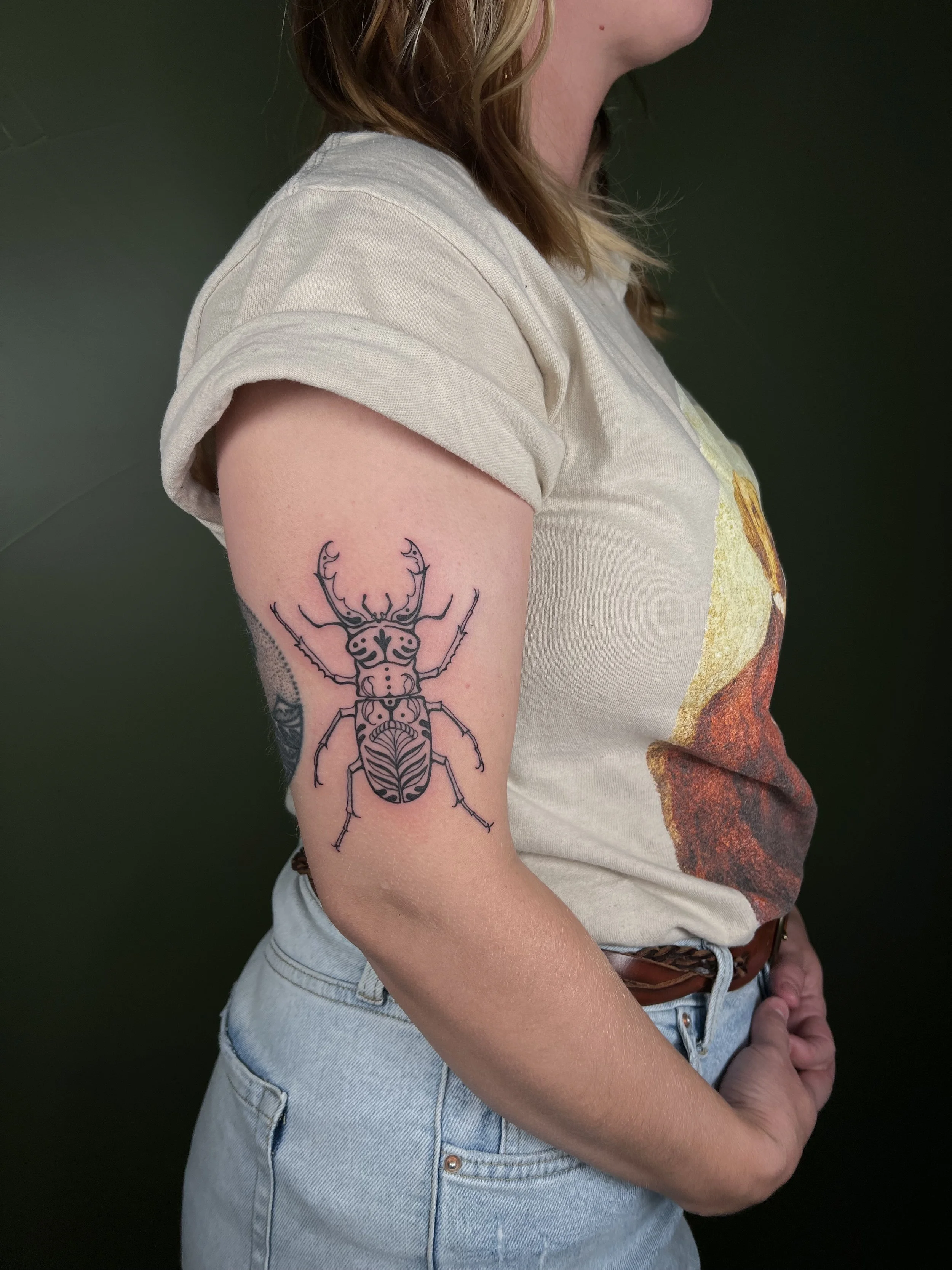 Tattoo of a beetle on a woman's upper arm, wearing a beige T-shirt with a graphic and light blue jeans against a dark background.