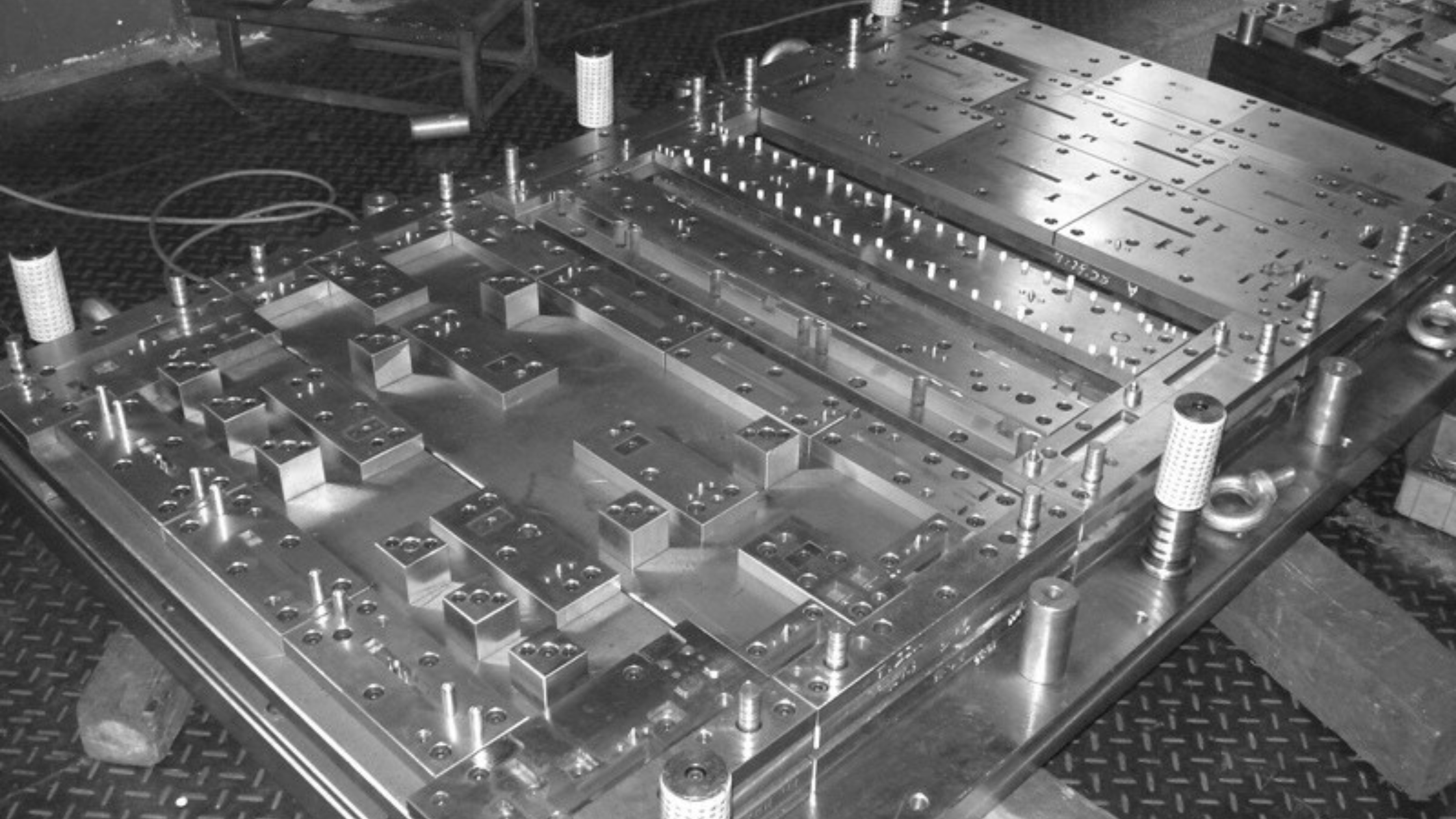 Close-up black and white photo of a metal mold with precise indentations and screws, set on a textured work surface with wires and other small tools nearby.