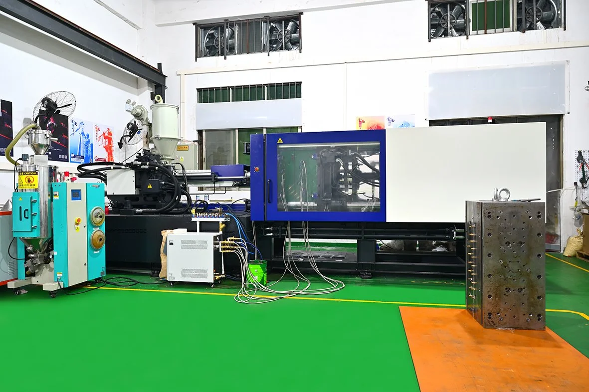 Industrial machinery in a workshop with green and orange flooring, featuring a large machine enclosed in a blue and white casing, connected to various wires and components.