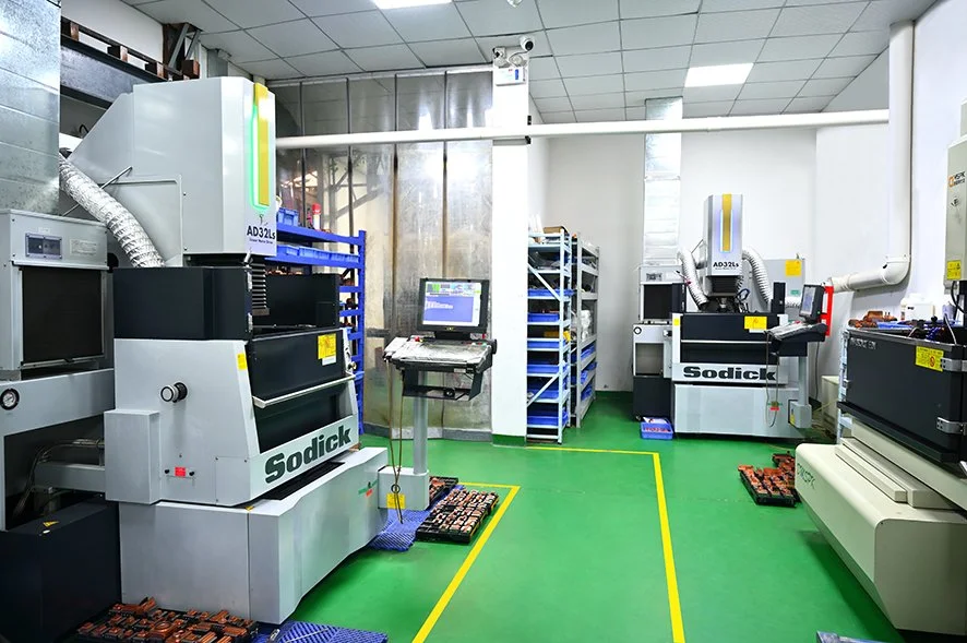 Industrial electronics manufacturing room with Sodick machines, computer workstation, storage racks, and safety markings on the green floor.