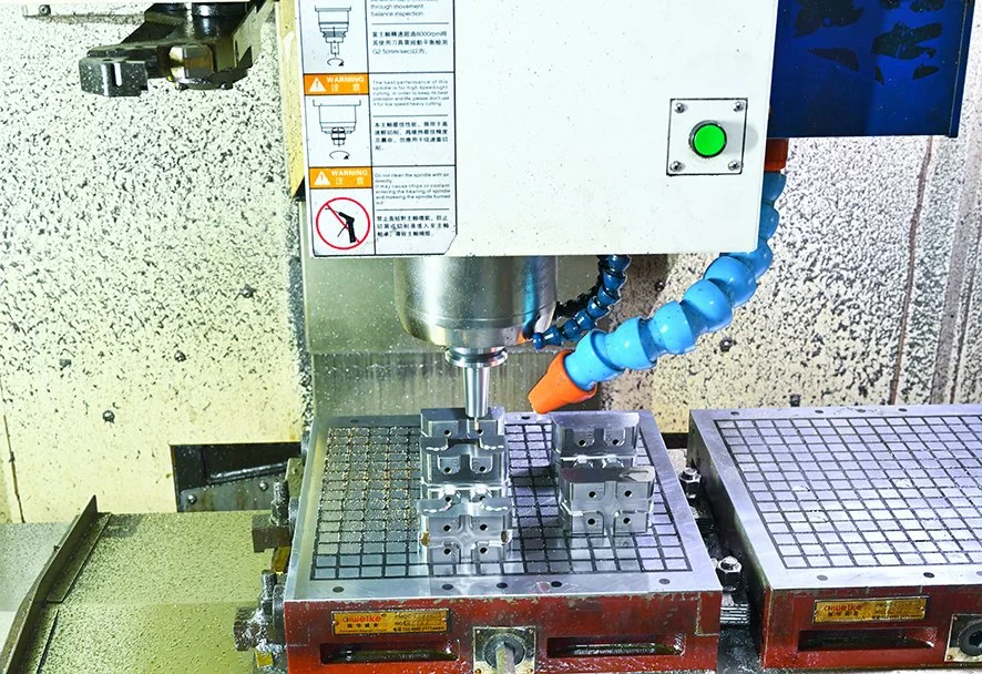 A CNC milling machine with metal blocks being machined. The machine has a vent tube and a green power button.