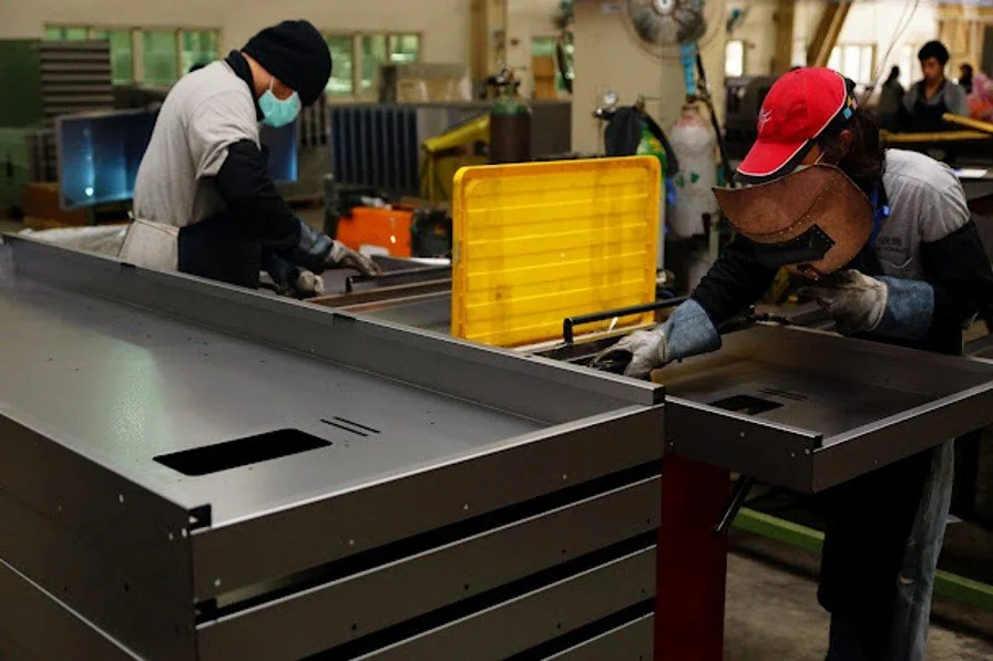 Two workers in a factory, both wearing protective gear and masks, assembling or inspecting metal parts on a large worktable.