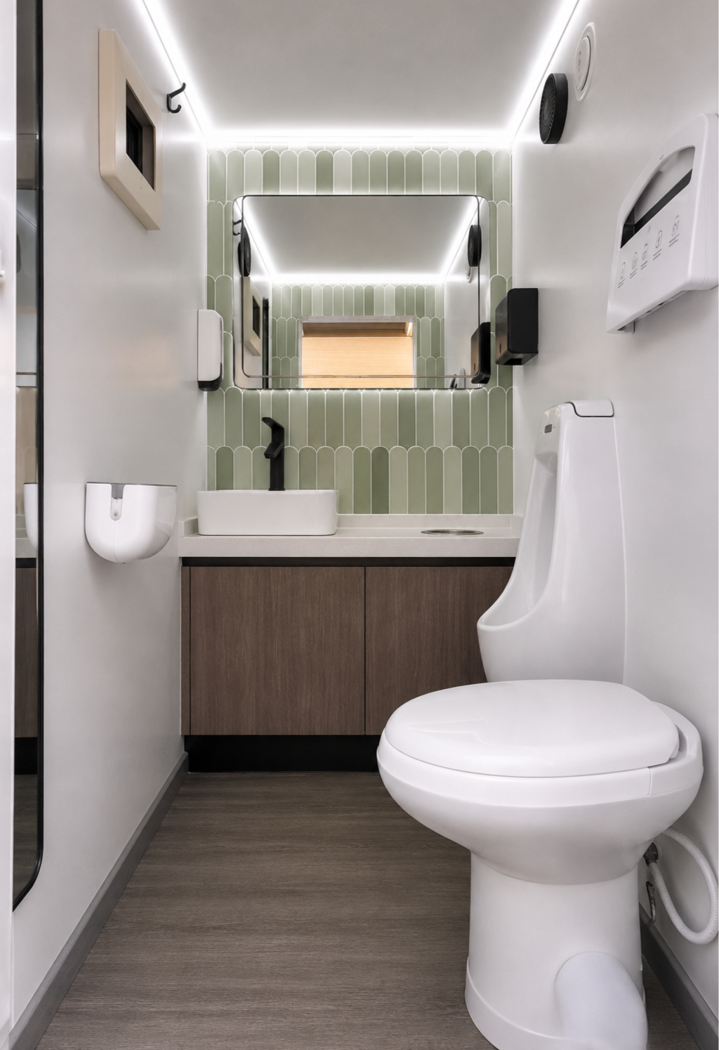 Small modern bathroom with white walls, green tile backsplash, a mirror, a black faucet sink, a wooden cabinet, a white toilet, a urinal, a wall-mounted soap dispenser, a mirror with LED lighting, and a wall-mounted speaker.