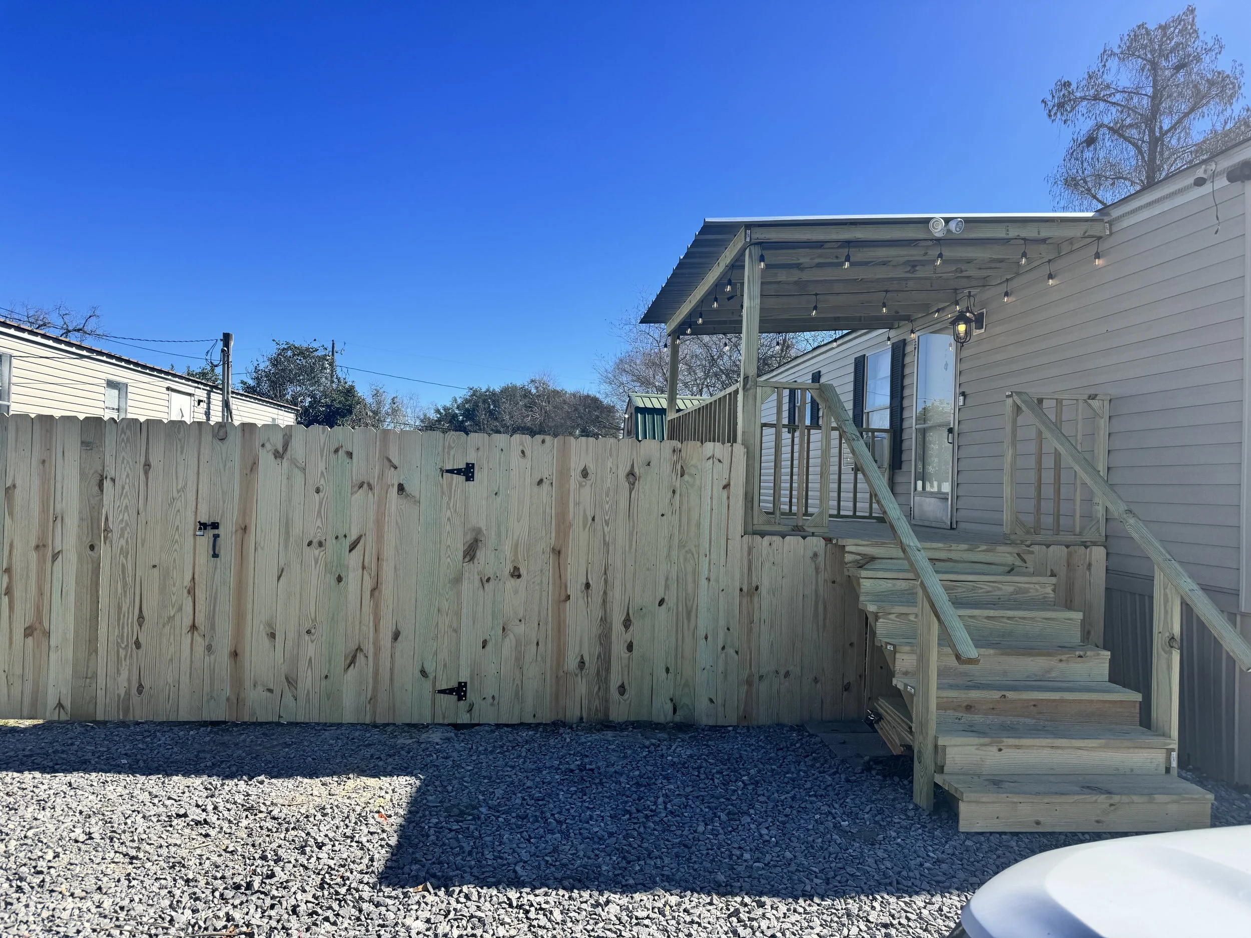 Backyard view showing a newly constructed wooden deck with stairs and railing, a partial fence, and string lights hanging from the deck's roof, with a gravel ground and a neighboring mobile home in the background.