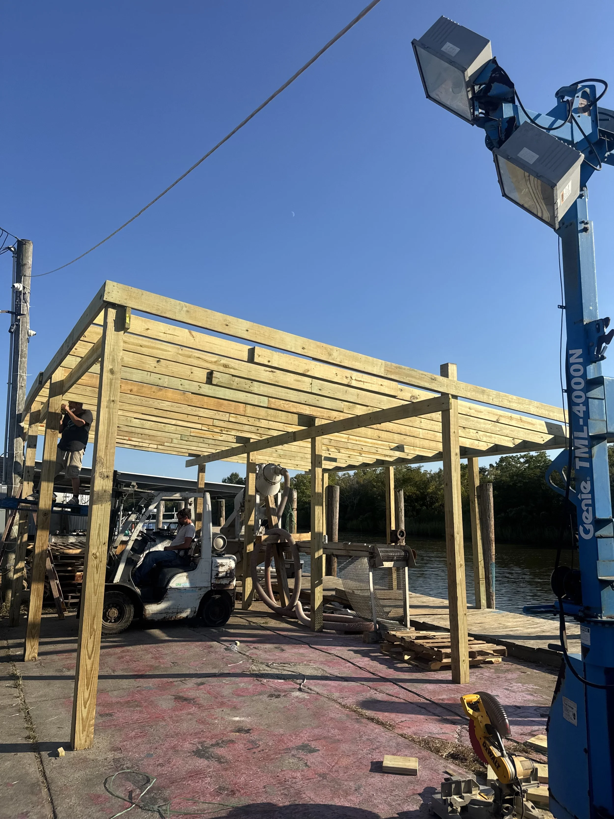 Construction site near water with workers building a wooden structure, a forklift, and bright blue sky.