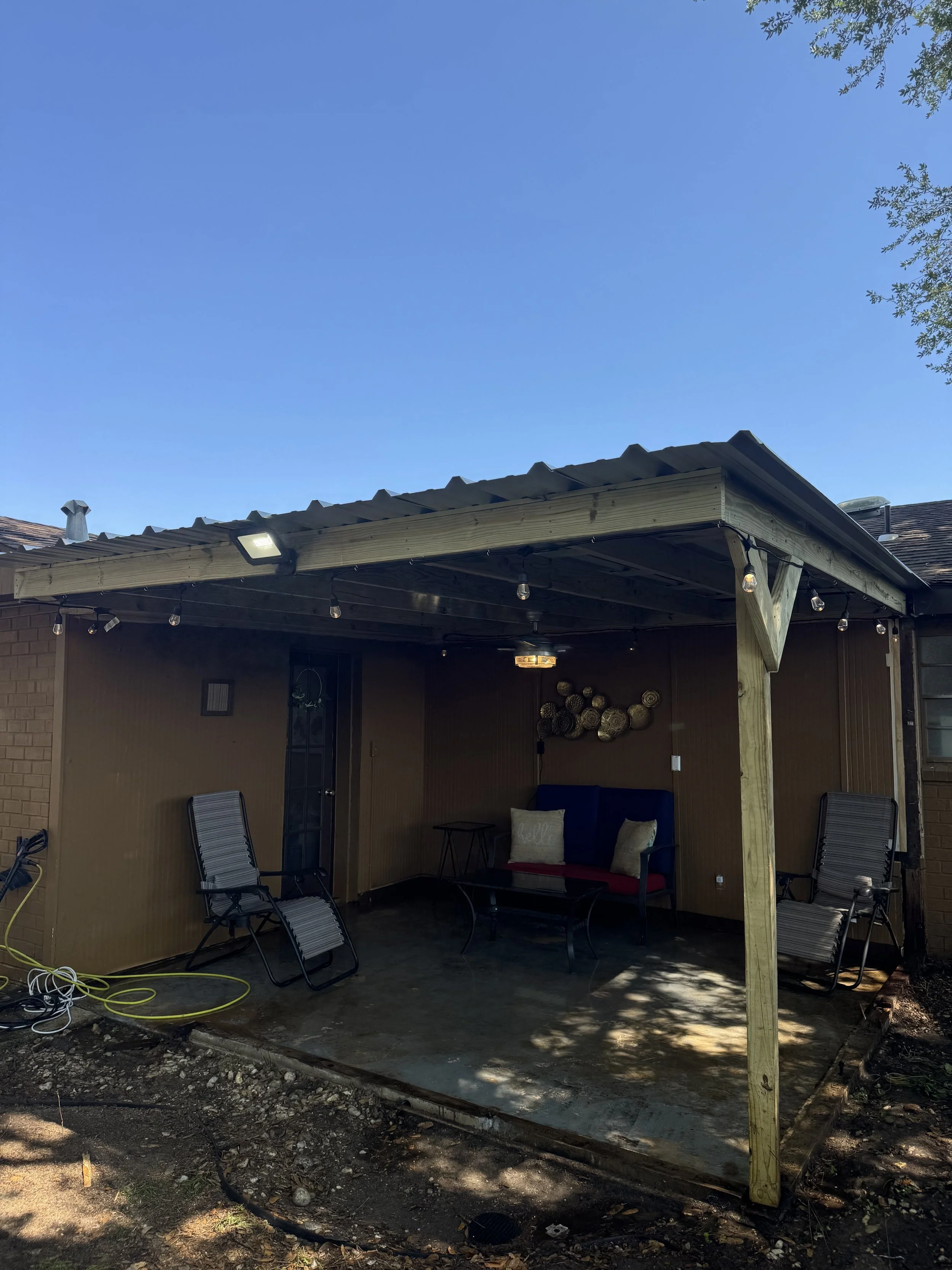 A backyard patio area with a small screened-in porch, furnished with a black metal cushioned bench, two lounge chairs, and a small table. String lights are hung along the porch roof, and decorative wall art is visible inside. The patio floor appears 