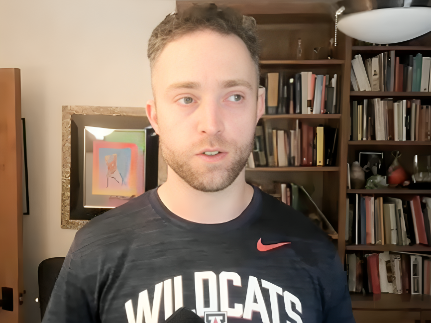 Young man with short, curly hair and a beard wearing a black T-shirt with 'WILDCATS' printed on it, standing in a room with bookshelves filled with books and decorative objects.