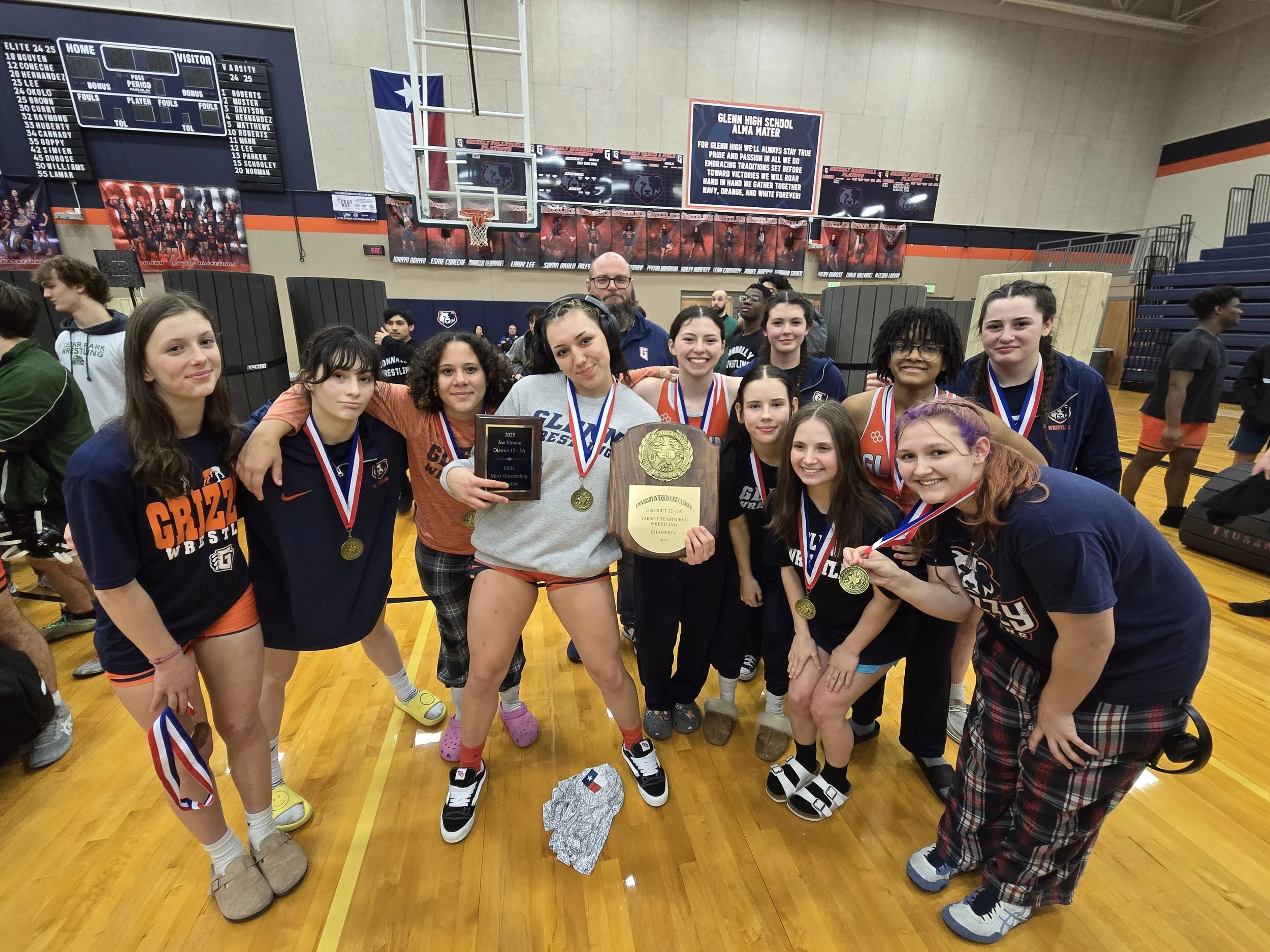 A group of young female wrestlers celebrating on a basketball court, holding medals and plaques, with a coach in the background. They are wearing sportswear with "Grizzlies" and "G" logos, and some have rainbow-colored hair and pajamas. The gym has banners and a scoreboard in the background.