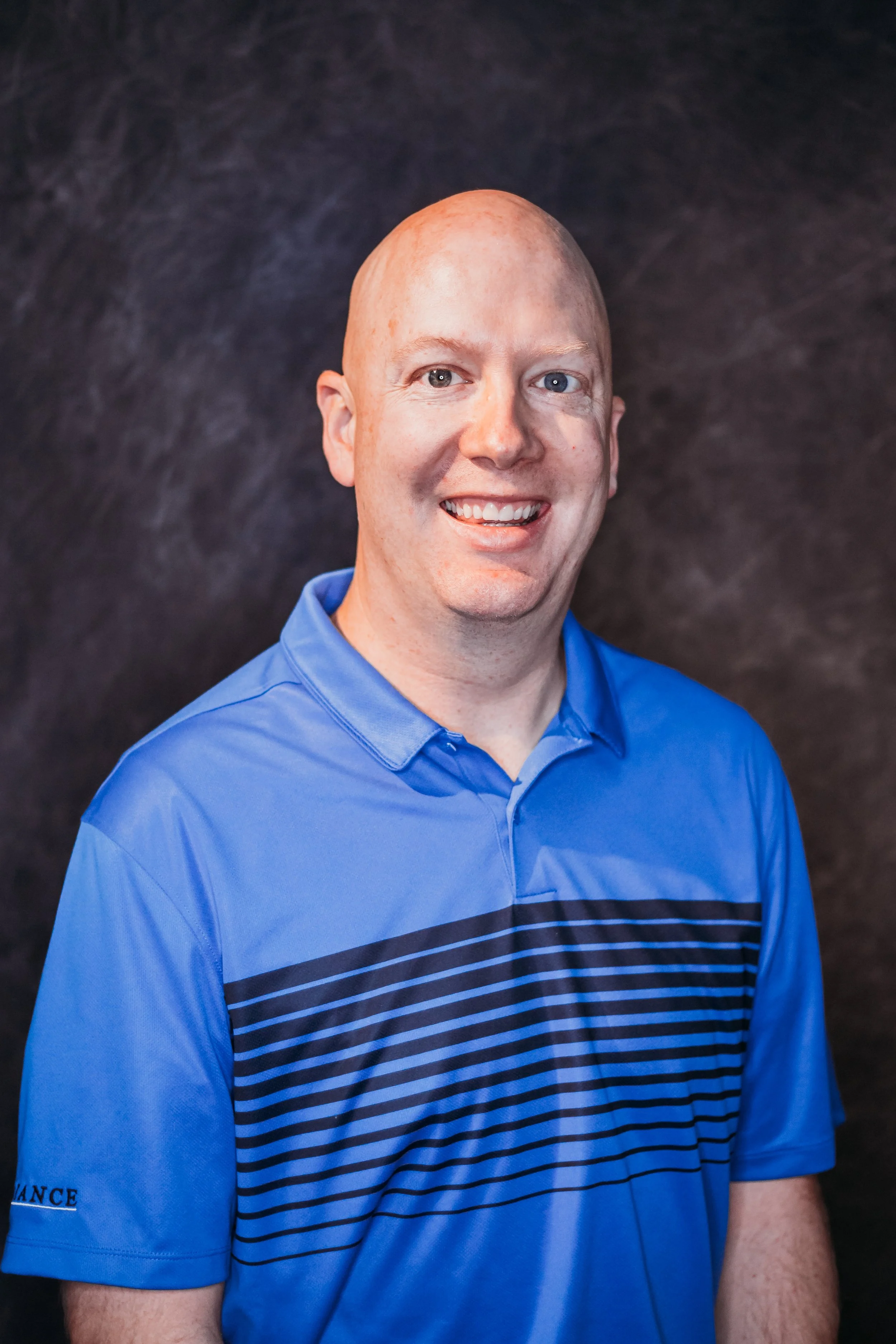 A smiling man with a bald head wearing a blue polo shirt with dark blue horizontal stripes, standing against a dark textured background.