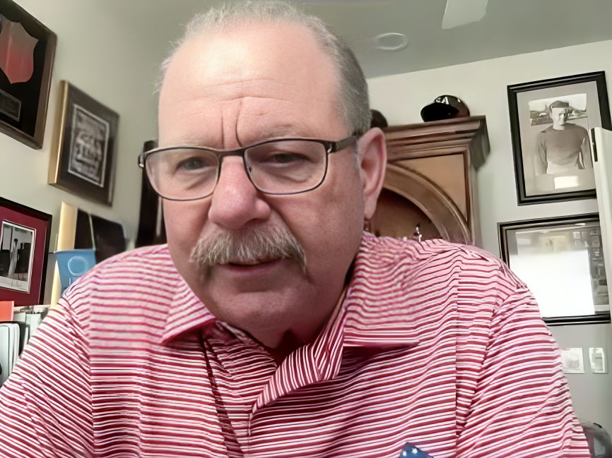Middle-aged man with glasses and a mustache wearing a red and white striped shirt, sitting in a room with framed photos and artwork on the walls.