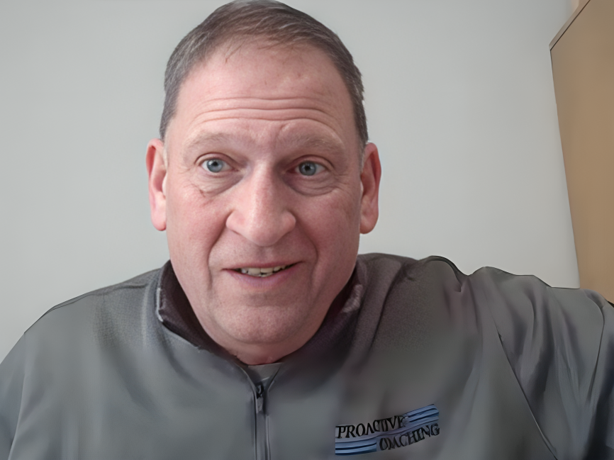 Close-up of a middle-aged man with short brown hair, blue eyes, and fair skin, smiling slightly while wearing a grey jacket with a logo that says 'Proactive Coaching'.