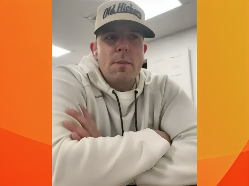 A young man wearing a white hoodie and a white baseball cap with black text, sitting indoors with arms crossed on a table.