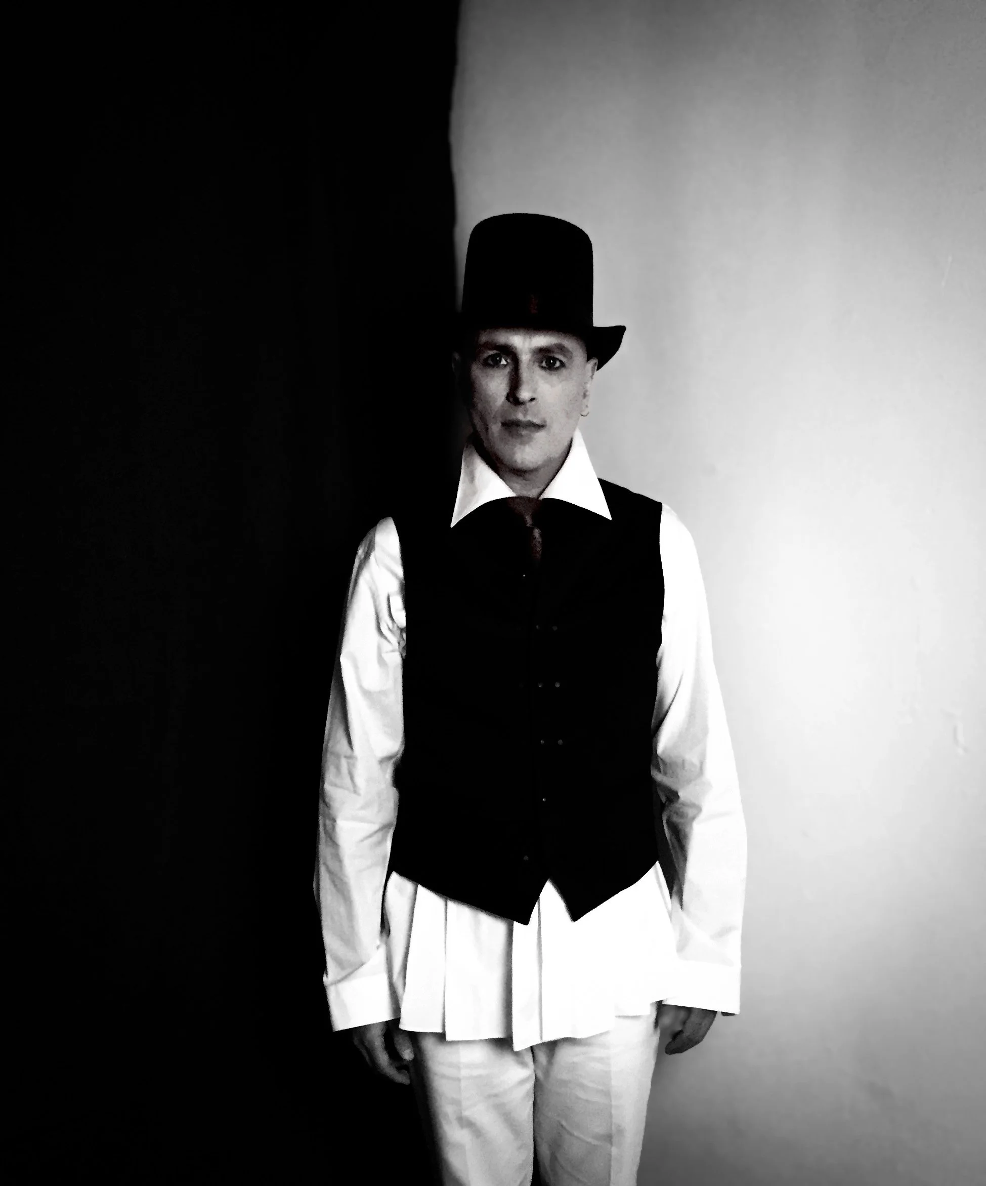JOHN DORIAN dressed in John Varvatos vest, Comme Des Garçons shirt, and tie, wearing a black top hat, standing against a background divided into black and white sections.
