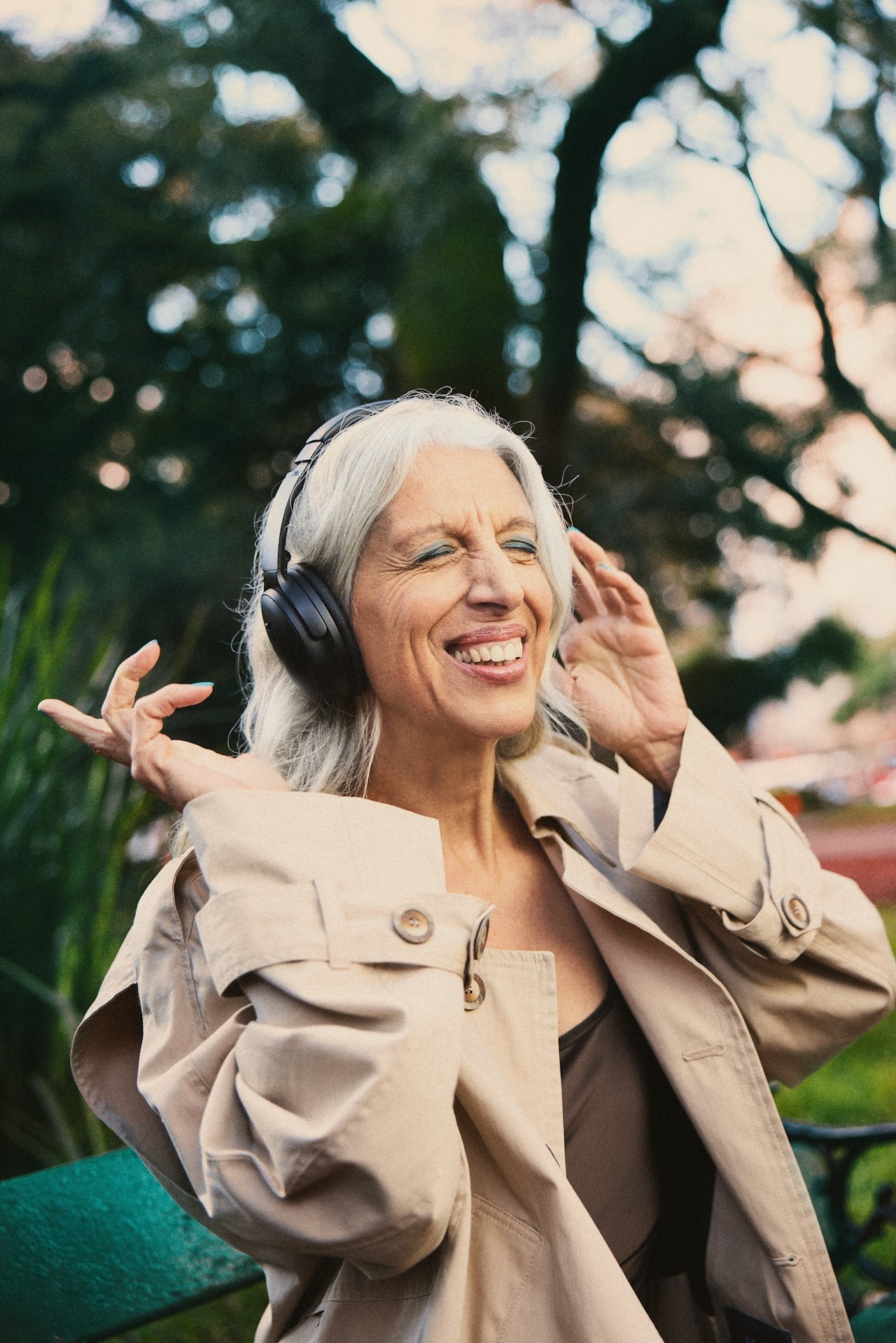 An elderly woman with gray hair, wearing a beige trench coat, is smiling with her eyes closed while listening to music on black headphones outdoors during the daytime.