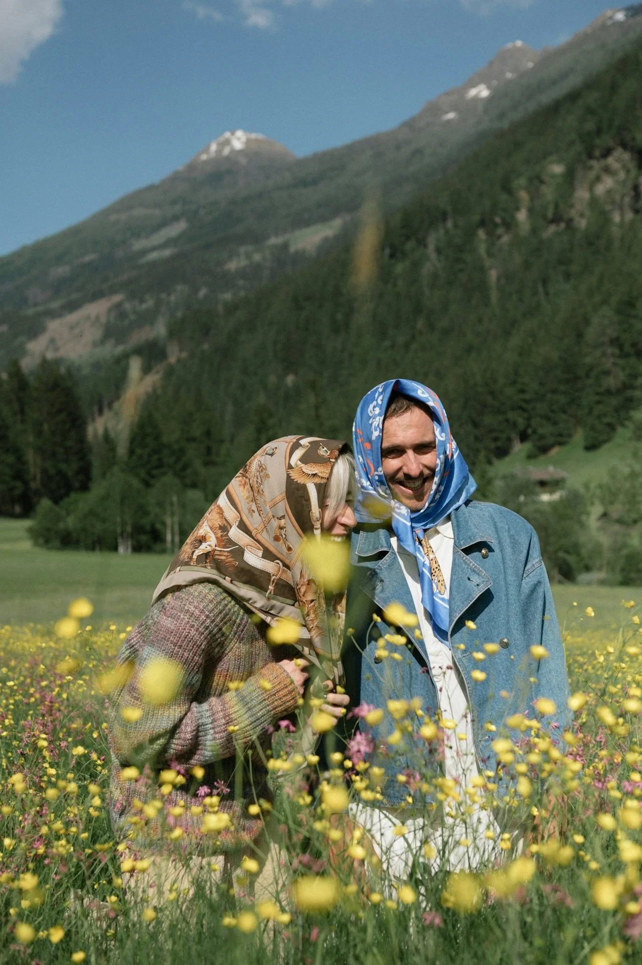 A smiling man and woman wearing colorful scarves on their heads, standing close together in a field of yellow and pink flowers, with green mountains and a blue sky in the background.