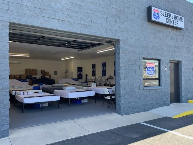 Interior of Sleep & Home Center with mattresses displayed and boxes on shelves, viewed through an open garage-style door outside a gray brick building.