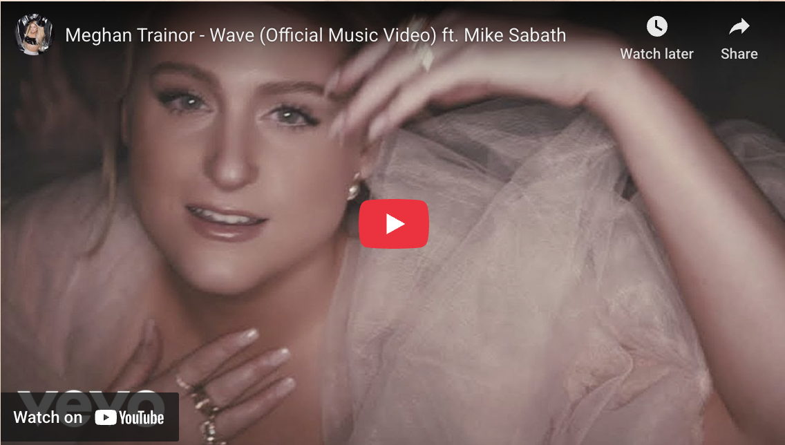 A woman with blonde hair and makeup, wearing a light-colored, sheer dress, lying down and smiling at the camera.