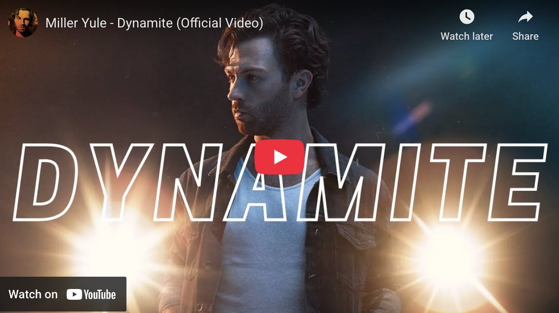 A man with dark curly hair and a beard, wearing a denim jacket over a white shirt, looking to his left with a serious expression. The background is dark with bright light flares behind him, and large white text says 'DYNAMITE' across the image.
