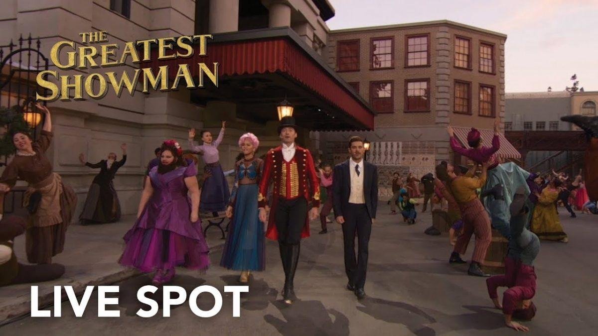 Musicians and performers dressed in colorful clothing dance and sing outside a building with large windows, while some are doing acrobatic stunts. Text overlay says "The Greatest Showman" and "Live Spot."
