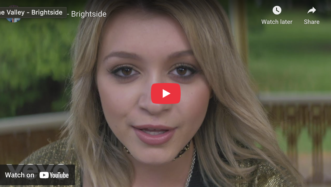 Close-up of a young woman with blonde hair and makeup, looking directly at the camera with a soft expression, outdoors with blurred greenery in the background.
