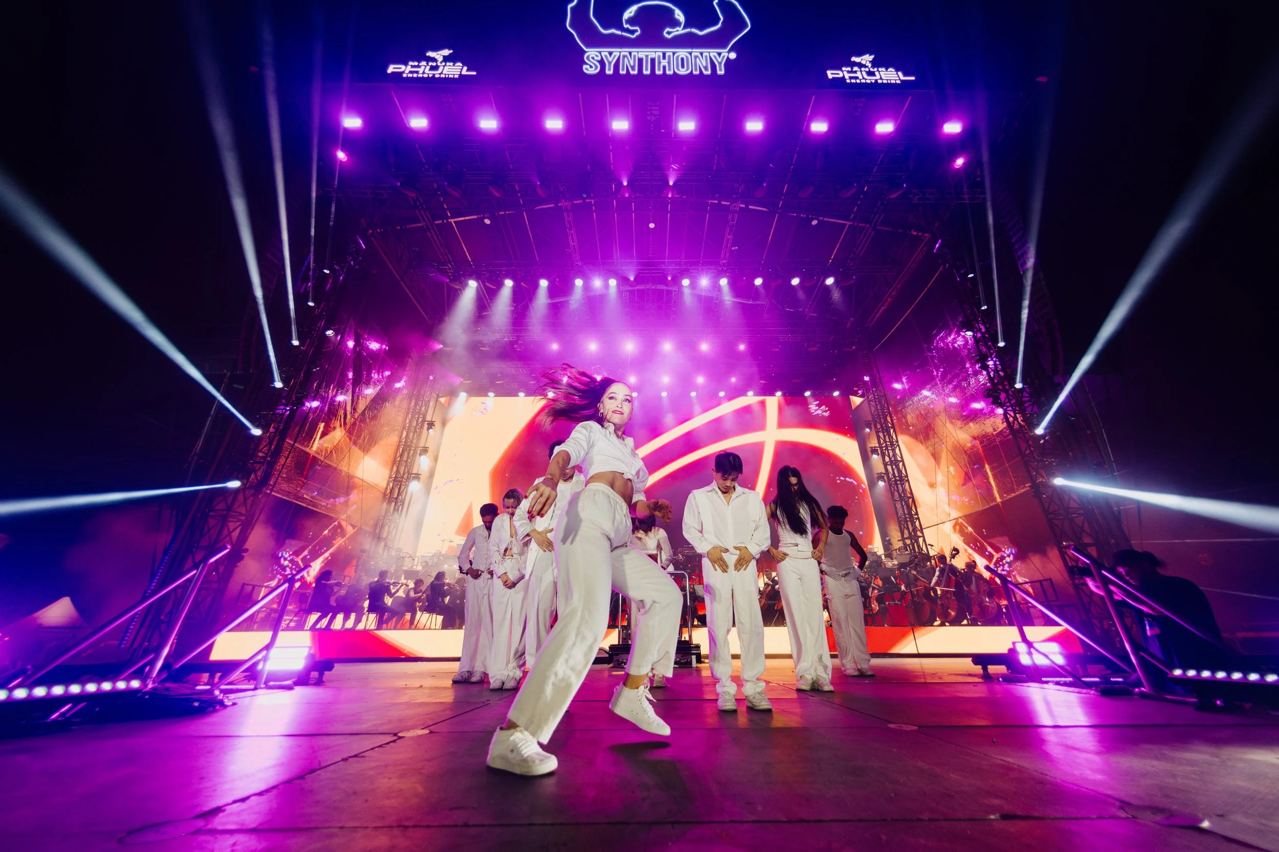 People dancing on stage during a concert with vibrant purple and orange lighting, a large digital screen showing abstract visuals, and a live orchestra in the background.
