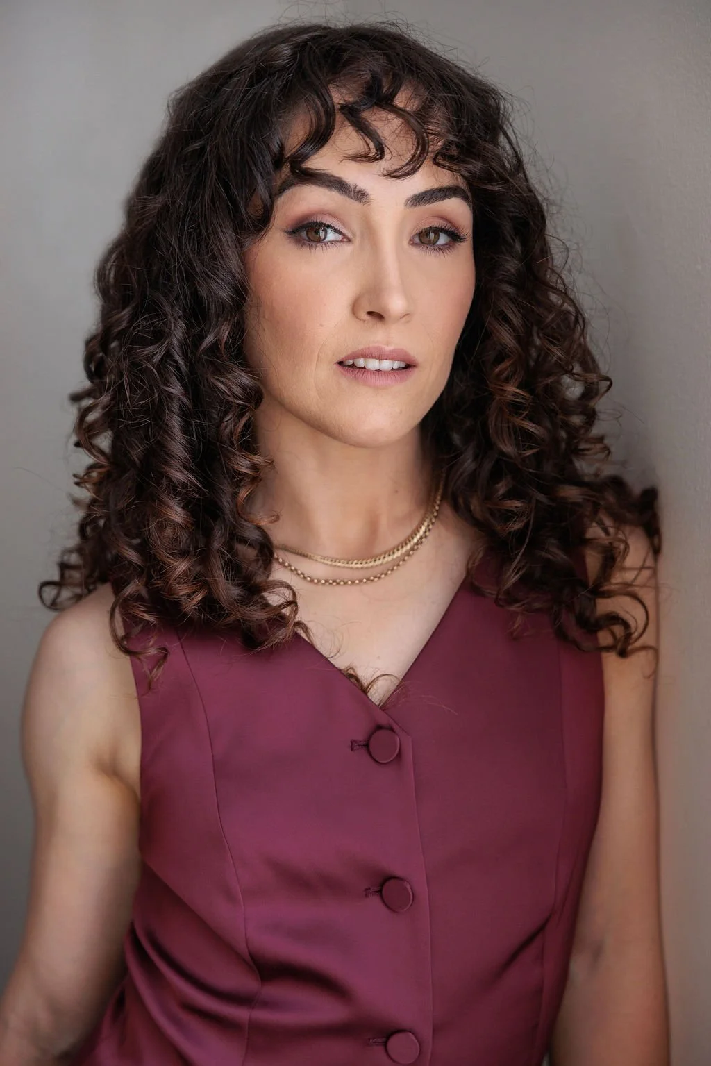 A woman with curly dark brown hair, wearing a sleeveless burgundy dress with buttons and layered gold necklaces, posing against a neutral background.