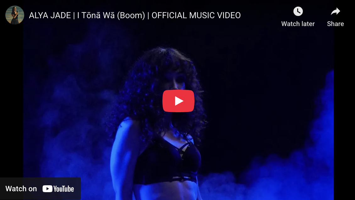 A woman with curly hair standing on a dark stage with blue lighting and fog, wearing a black outfit, during a music video performance.