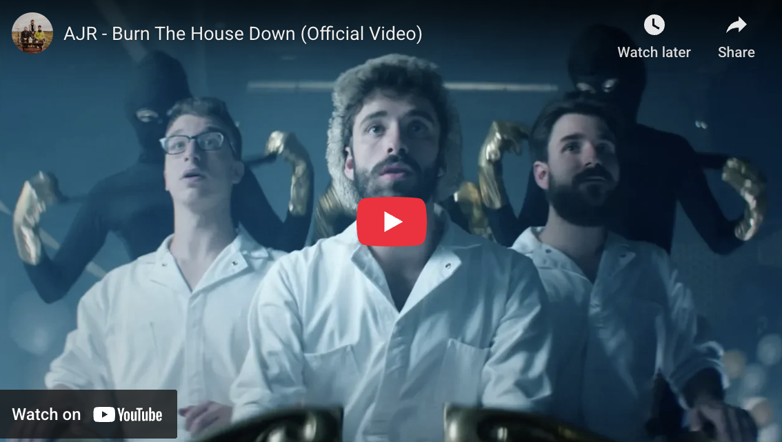 Four men in white lab coats are in the foreground, with two more shadowy figures behind them, wearing masks and black suits. The background is dark and blue-toned. This scene is from the music video "Burn The House Down" by AJR.