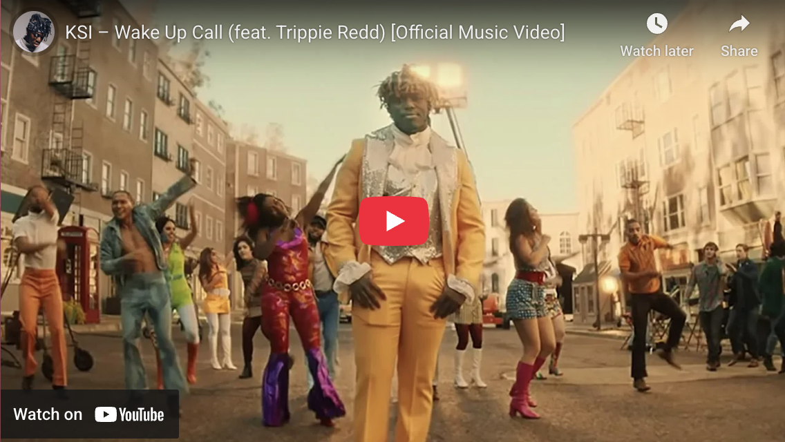 A music video scene with a man in a yellow suit standing in the center, surrounded by dancers in colorful outfits dancing on a city street with apartment buildings and a fire in the background.