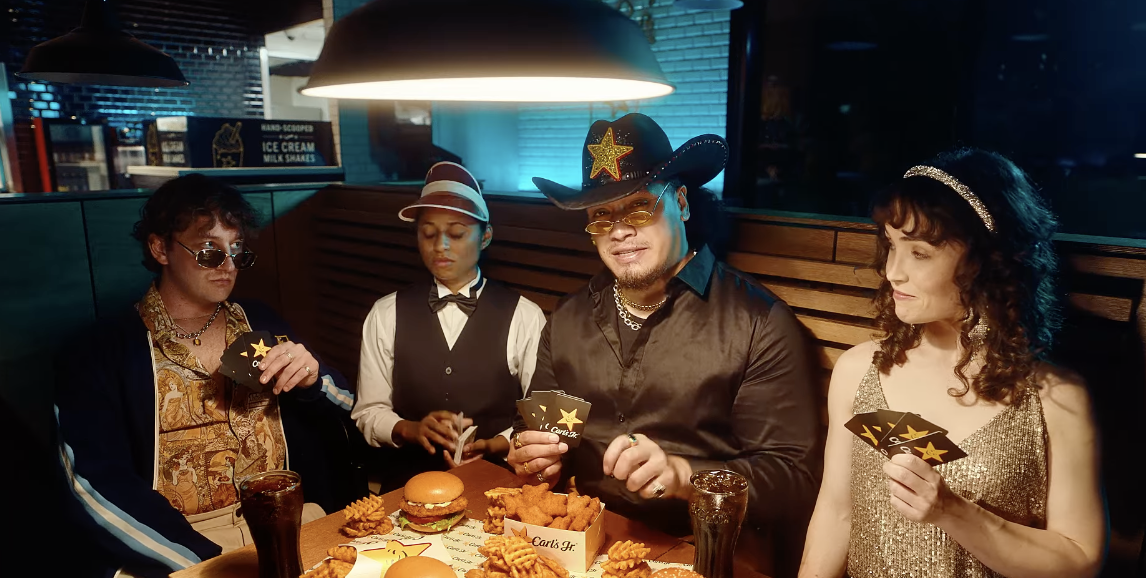 Four people sitting at a table in a restaurant playing cards and enjoying burgers and fries, dressed in festive costumes and accessories.