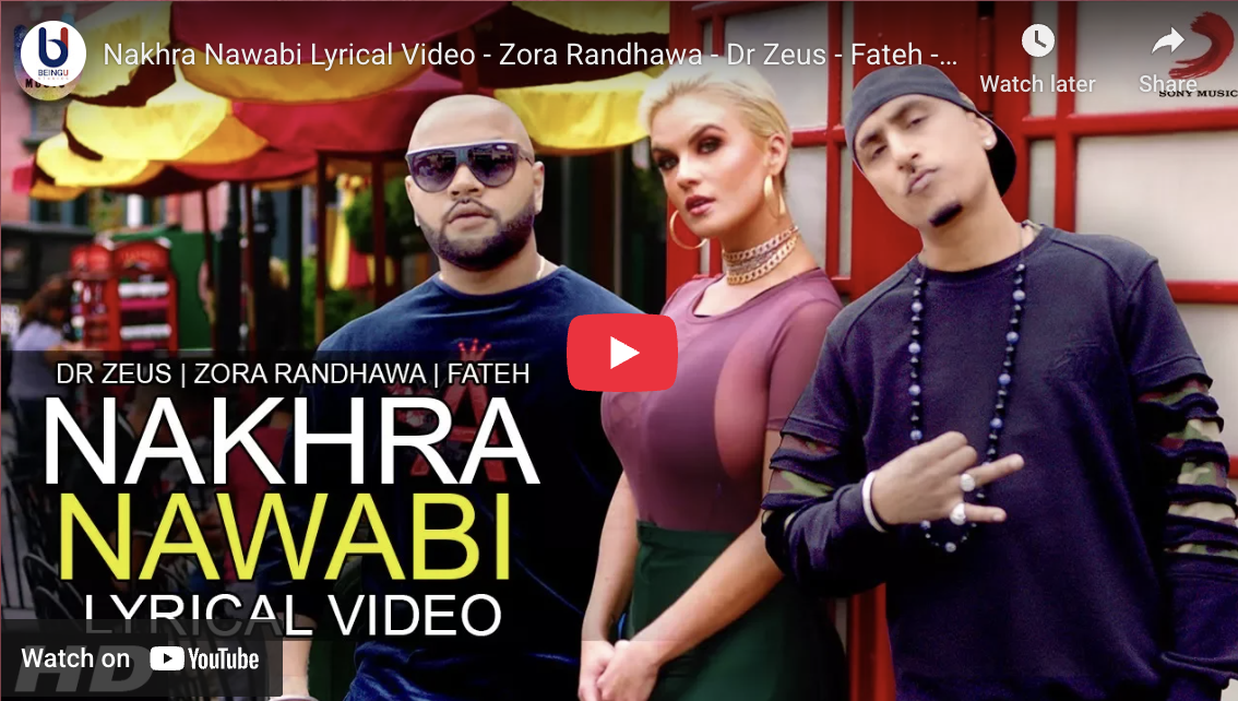 Three people standing outdoors in front of colorful hanging umbrellas, posing for a photo. The person on the left is wearing sunglasses and a navy blue shirt, the woman in the middle has blonde hair and is wearing a maroon top with gold jewelry, and 