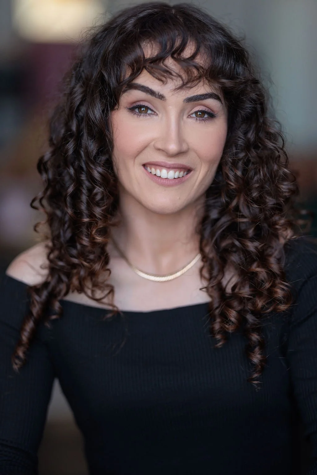 A woman with curly brown hair, hazel eyes, wearing a black top and a gold necklace, smiling.