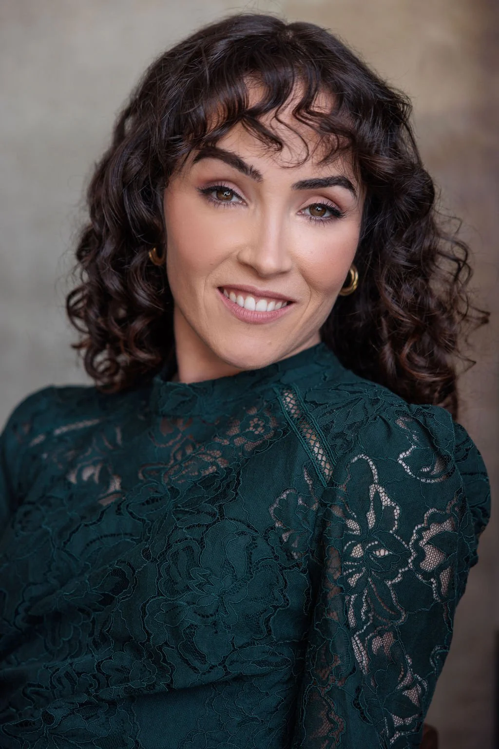 A woman with dark, curly hair, wearing a dark green lace top, smiling at the camera.