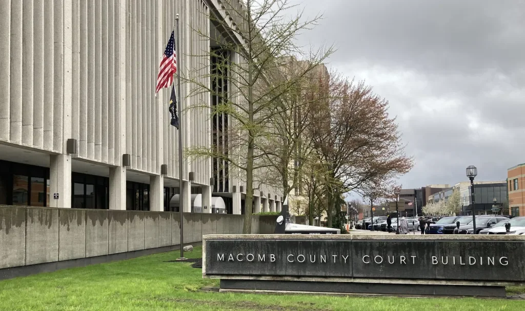 Exterior of Macomb County Court Building with American and state flags, trees, and parking lot.