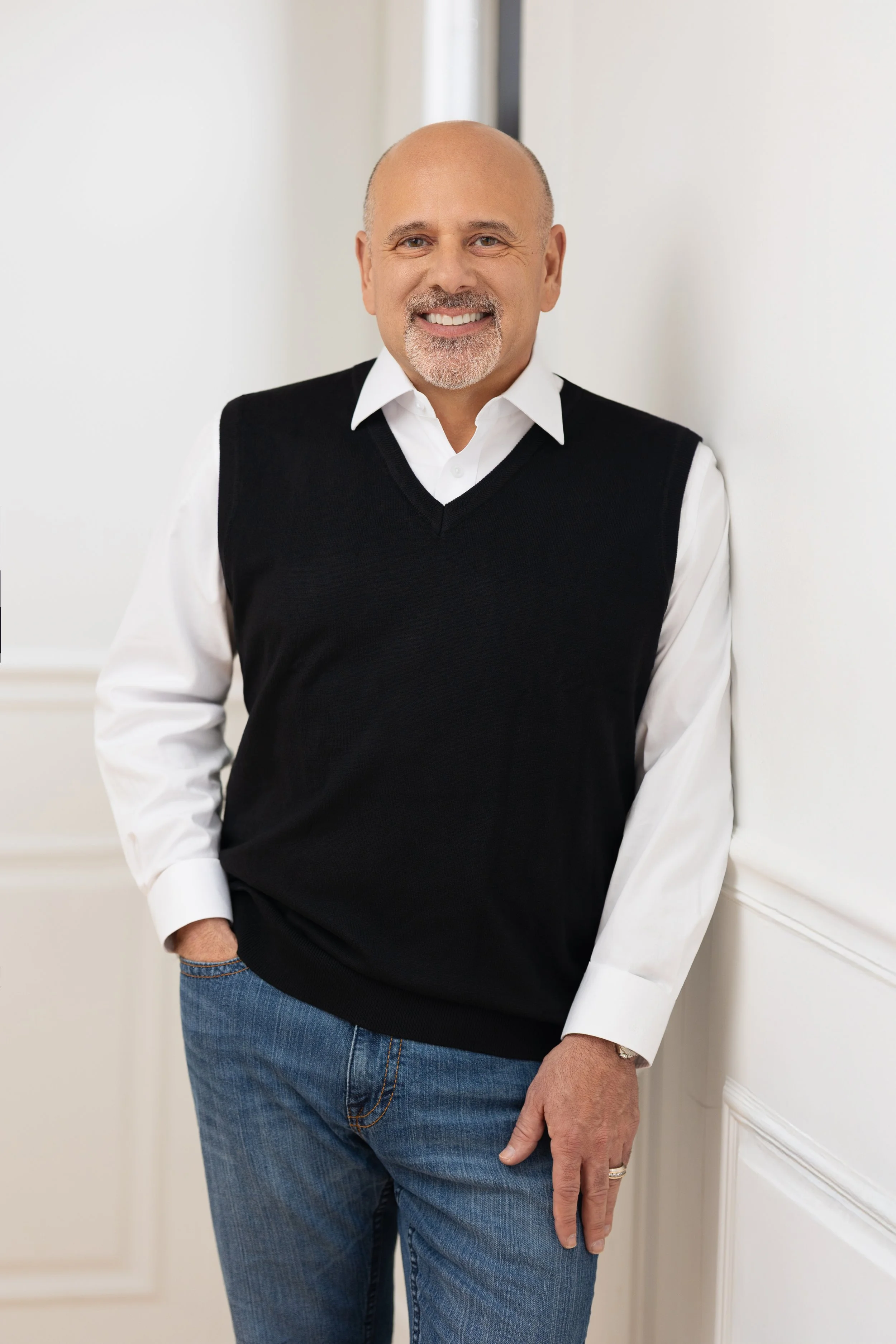 A smiling middle-aged man with a bald head and grey beard, wearing a white dress shirt and a black sweater vest, standing in front of a white wall.