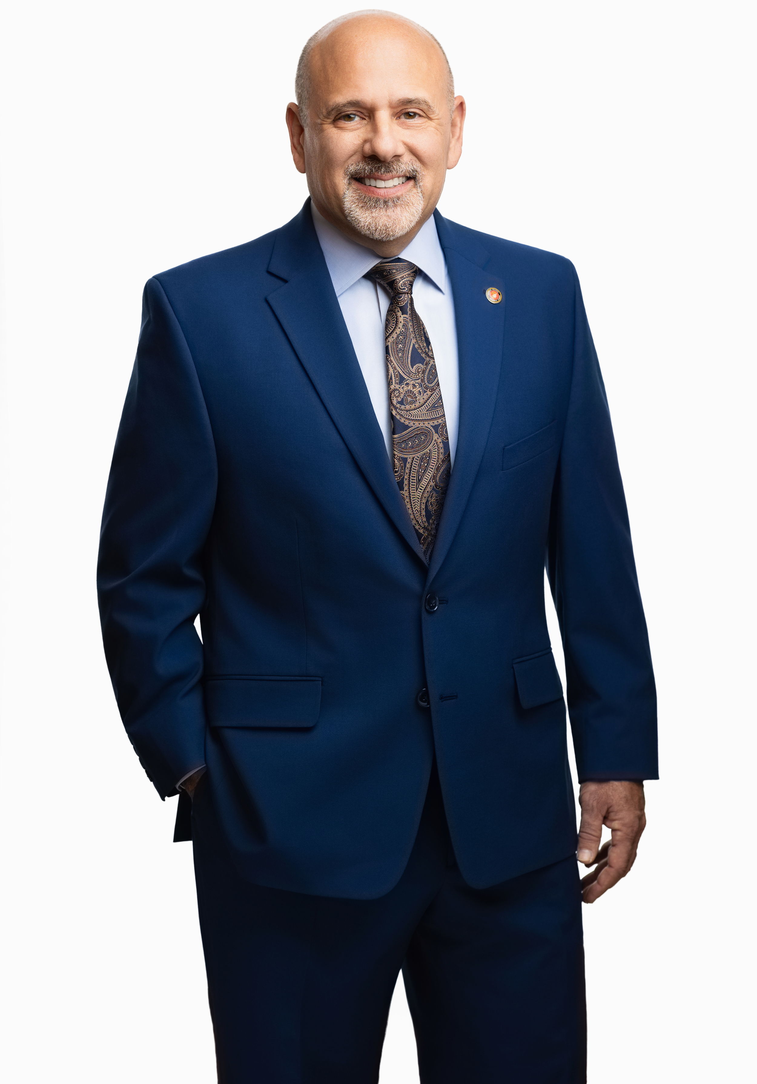 A smiling middle-aged bald man with a goatee, wearing a dark blue suit with a paisley tie, standing against a plain white background.