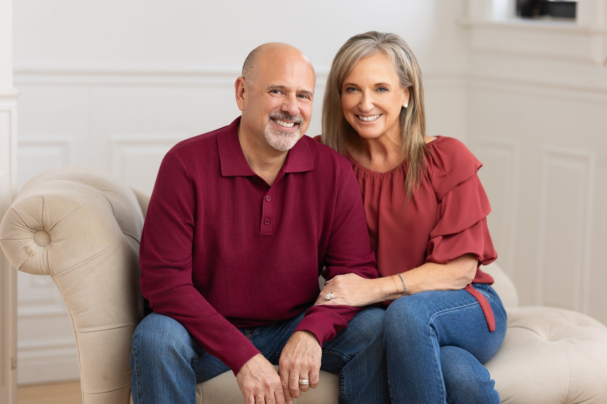 Smiling middle-aged couple sitting on a beige couch in a well-lit room, with the woman holding the man's hand and both dressed casually.