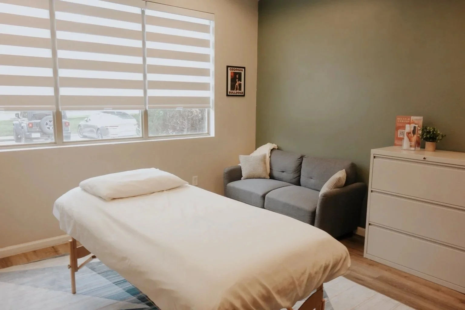 Private treatment room with a massage table, couch seating, dresser storage, and large windows.