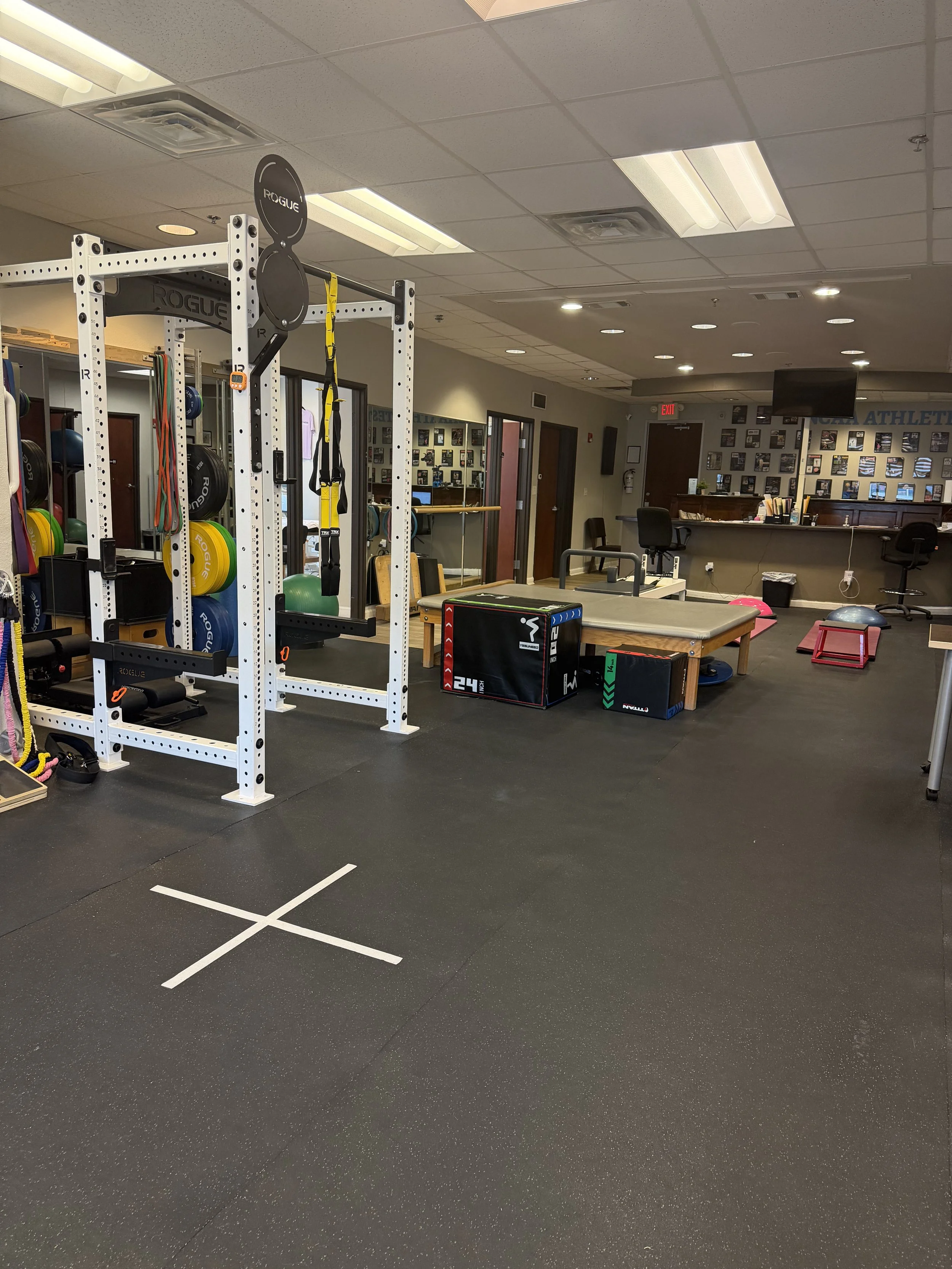 Empty gym or fitness training area with workout equipment, mats, weights, and a reception desk in the background.