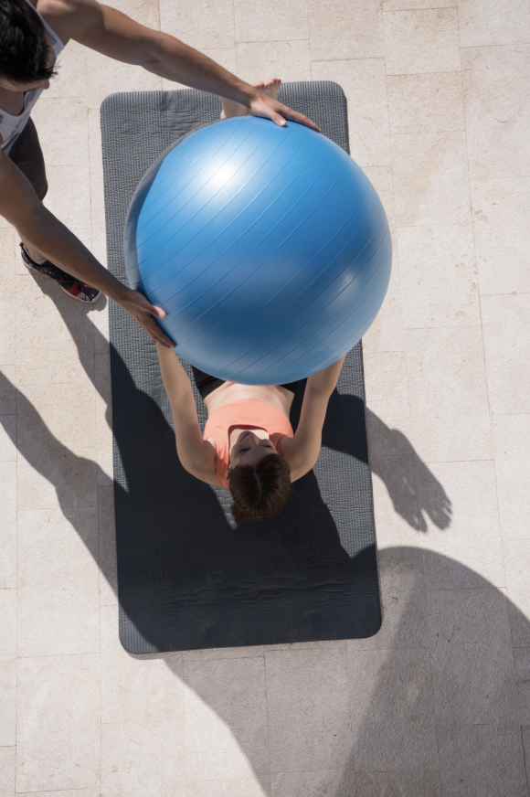 A person lying on a black exercise mat outdoors, holding a large blue exercise ball with assistance from another person.