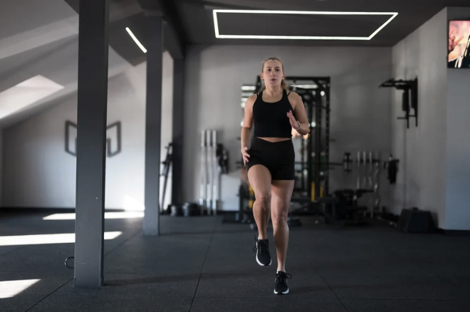 A woman running indoors at a gym, wearing a black sports top and shorts, with gym equipment in the background.