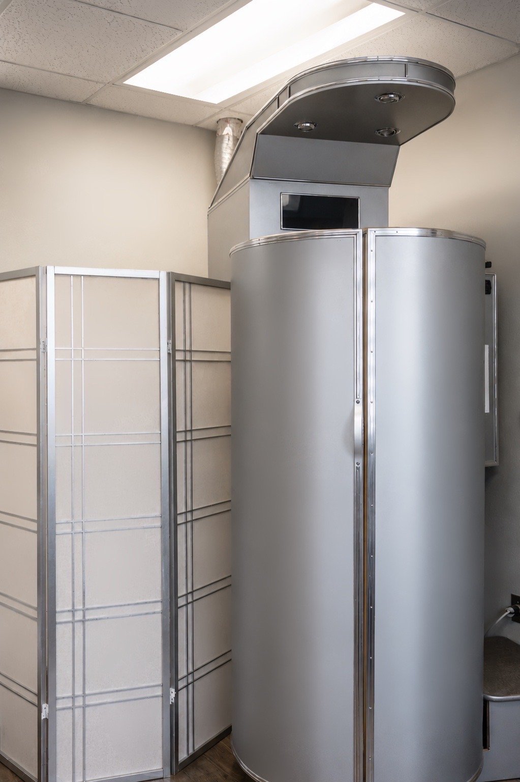 A silver cryotherapy machine with a monitor on top and a control panel on the side, located in a medical or therapy room.
