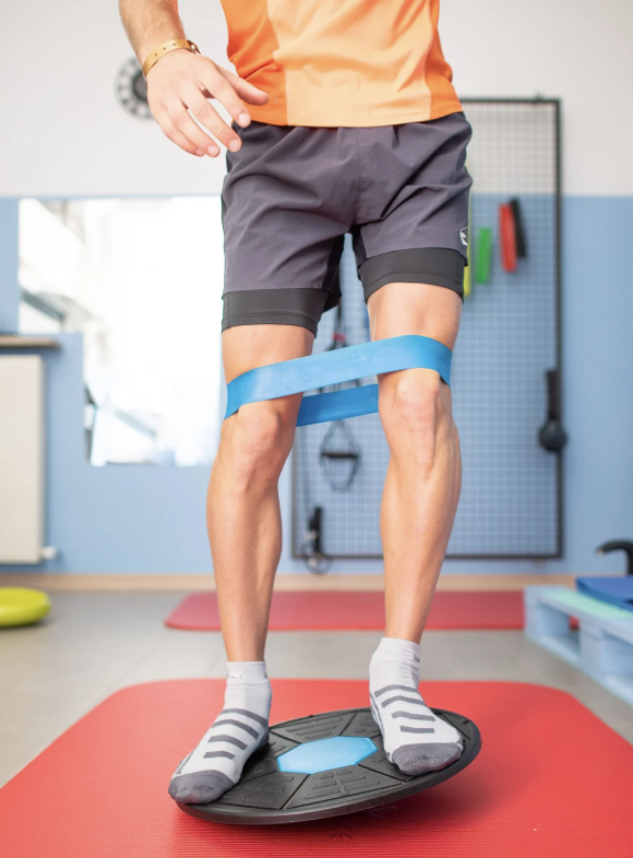 Person standing on a balance board with a blue resistance band around their thighs, performing physical therapy or balance training in a fitness room.