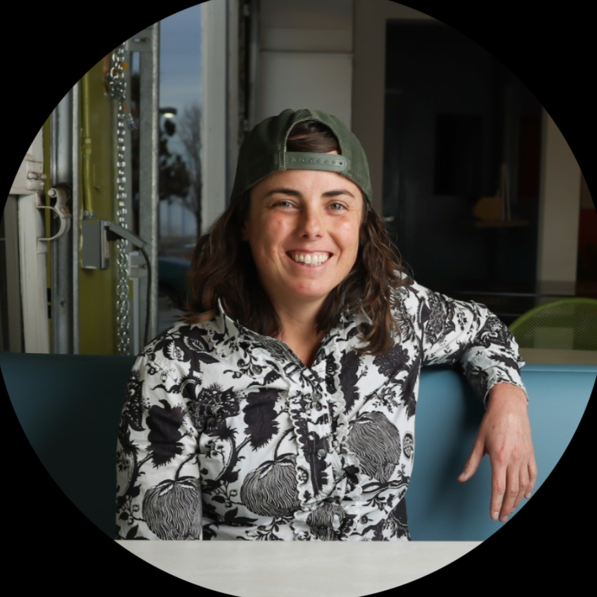Woman with dark wavy hair wearing a green baseball cap backwards and a black-and-white patterned shirt, smiling, sitting at a table in a modern indoor setting.