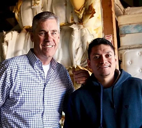 Two men are smiling and posing together in a warehouse or storage area with large bags or sacks in the background.