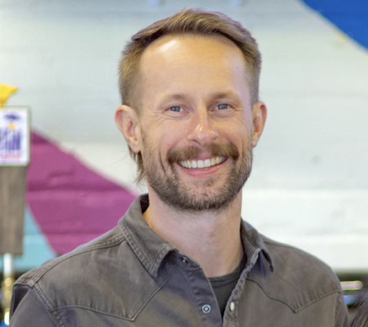 A smiling man with a beard and short, light brown hair, wearing a gray button-up shirt, standing indoors with colorful decor in the background.