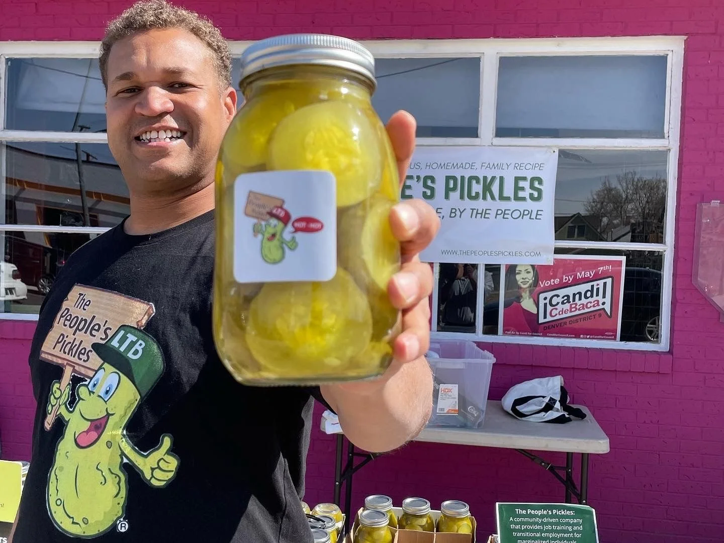 Smiling man holding jar of pickles outside pink building with signs for The People's Pickles and vote campaign in the background.
