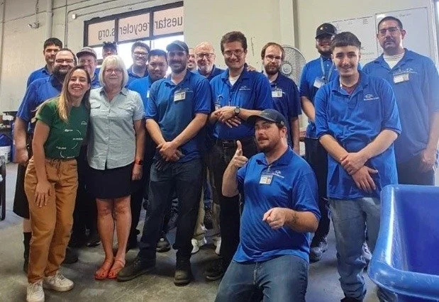 Group of people, some wearing blue uniforms, standing in a room with a sign that says 'Useful [illegible]'. They are smiling, posing for a group photo, with one person kneeling in front giving a thumbs-up.