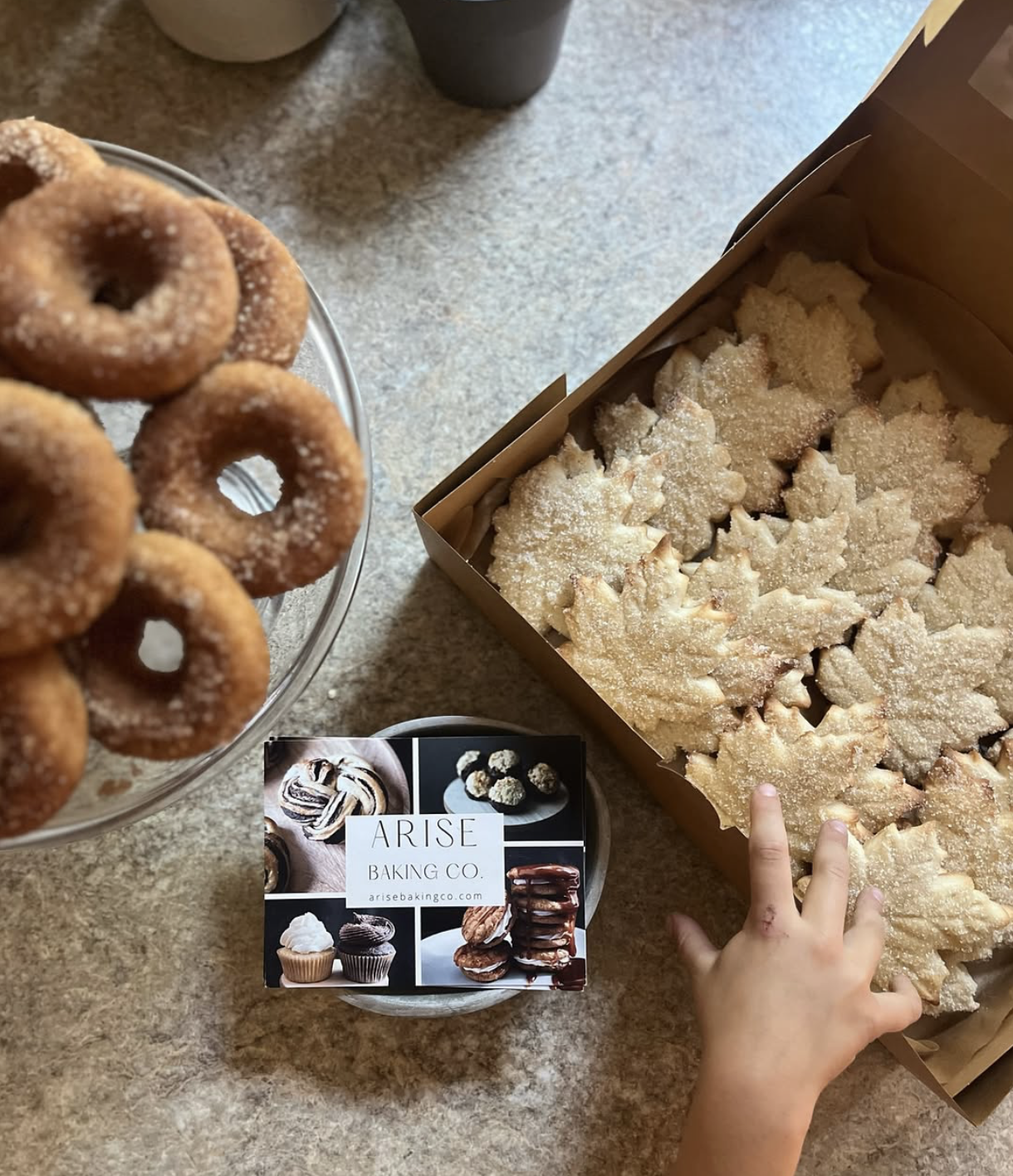 A toddler's hand reaching toward a box of sugar-coated cookies on a surface. To the left, a glass stand holds a stack of churros with sugar on top. In the center, there is a small round container with a collage of bakery images and the label 'Arise B