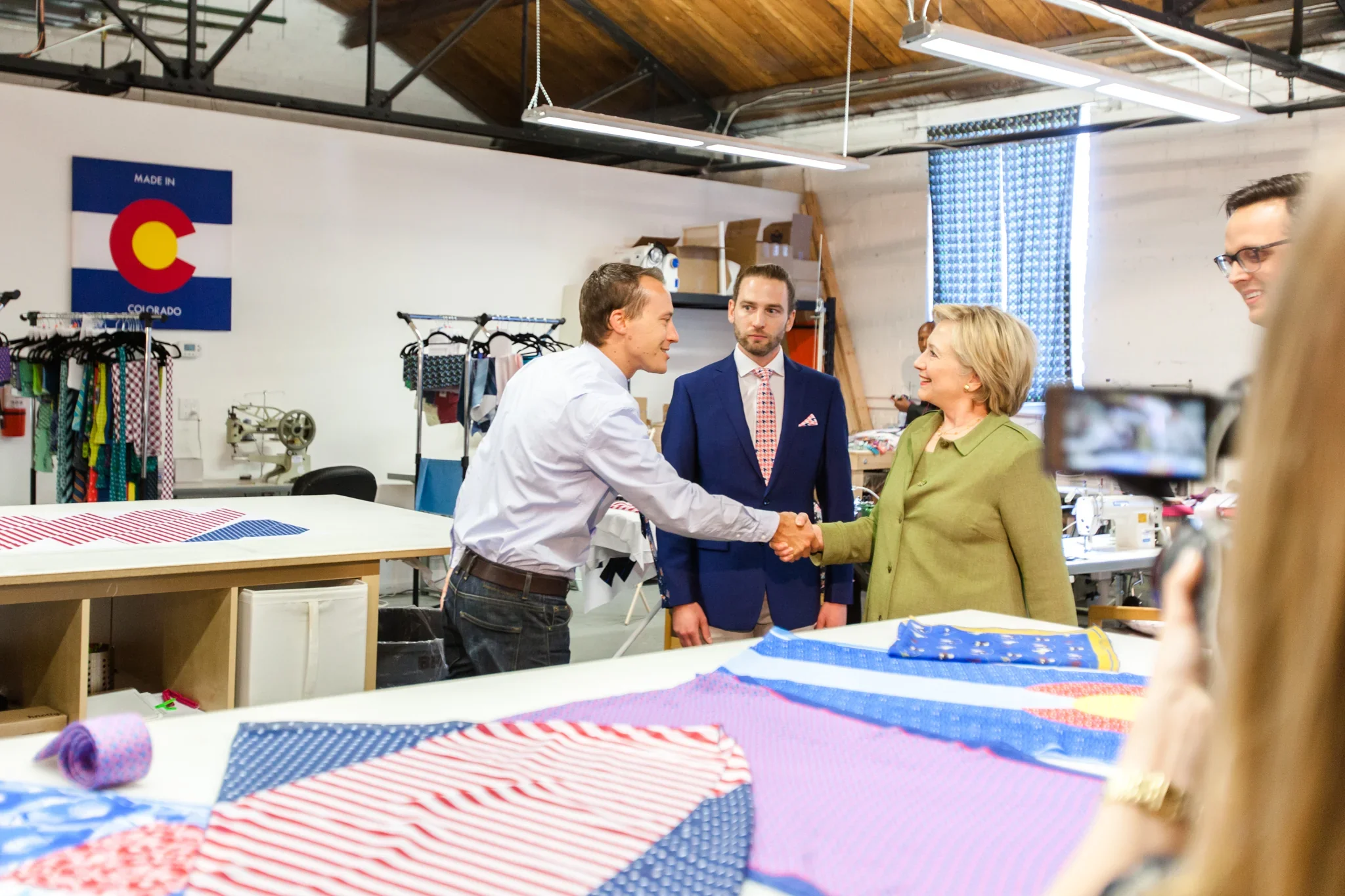 A man in a white shirt shaking hands with a woman in a green jacket, standing in a fabric or clothing workshop. Two men in suits observe the handshake, with the workshop filled with fabric, sewing machines, and a Colorado flag on the wall.